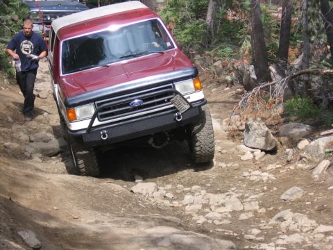 1989 Ford Bronco - A red Ford Bronco navigates a challenging, rocky off-road trail with a person walking alongside.