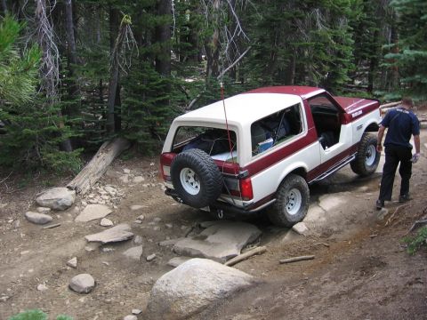 1989 Ford Bronco - A two-tone red and white Ford Bronco navigates a challenging, rocky trail through a forest.