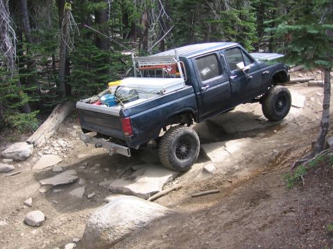1989 Ford Bronco - A lifted pickup truck navigates a rocky off-road trail in a wooded area.