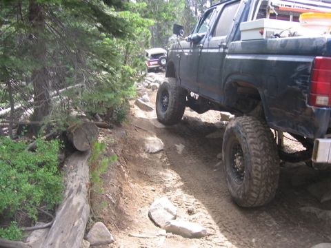 1989 Ford Bronco - Two modified trucks navigate a challenging off-road trail through a wooded area.