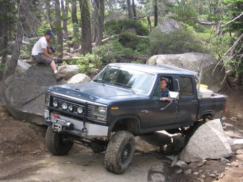 1989 Ford Bronco - A man drives a lifted Ford pickup truck over rocks on a wooded trail.