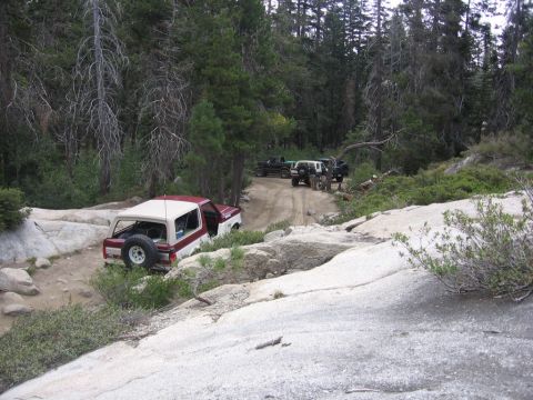 1989 Ford Bronco - A red and white 1989 Ford Bronco is parked off-road, with several other SUVs visible on a dirt path in the background.