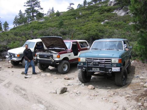 1989 Ford Bronco - Three Ford Broncos are parked on a dirt road, with the teal Bronco in the foreground a winch and custom bumper.