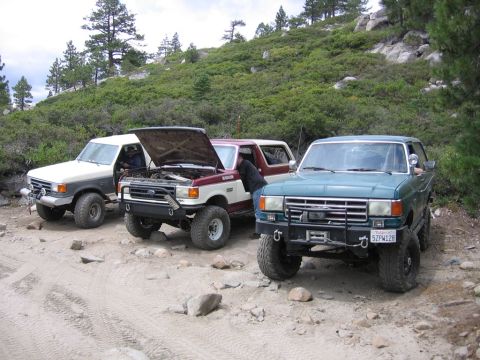 1989 Ford Bronco - Three Ford Bronco SUVs are parked on a rocky dirt road, with a wooded hillside in the background. One of the vehicles has its hood open.