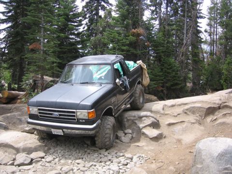 1989 Ford Bronco - A 1989 Ford Bronco navigates a rocky off-road trail surrounded by trees.
