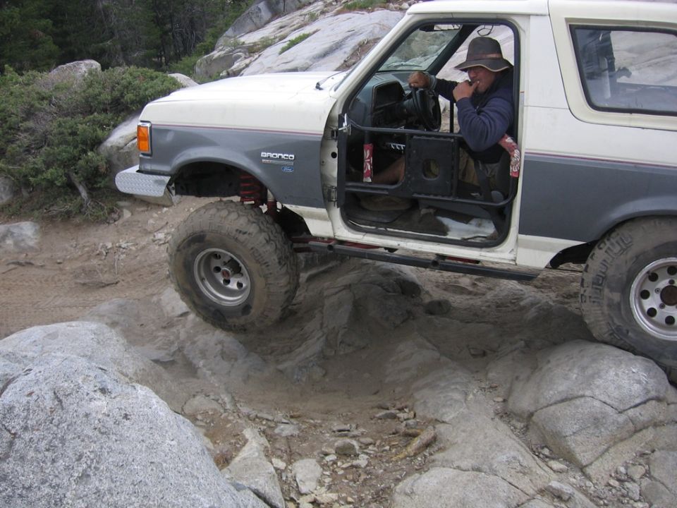 White and grey 1989 Ford Bronco off-roading over rocks, driver wearing a hat