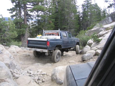 1989 Ford Bronco - A dark blue Ford F-Series pickup truck navigates a challenging off-road trail with rocky terrain and trees.