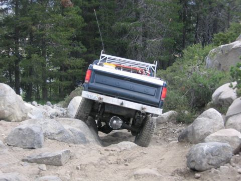 1989 Ford Bronco - A dark blue Ford pickup truck is shown navigating a challenging rocky trail, viewed from the rear.