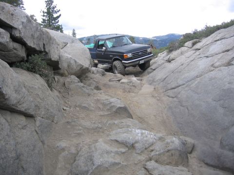 1989 Ford Bronco - A dark-colored Ford truck is shown driving over a rough, rocky terrain with large boulders on either side.