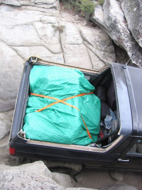 1989 Ford Bronco - A vehicle is wedged between rocks, its cargo area covered with a green tarp and secured with orange straps.