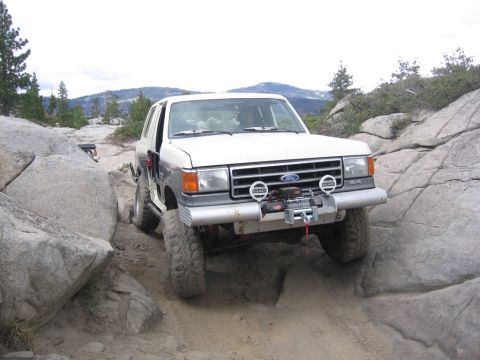 1989 Ford Bronco - A white 1989 Ford Bronco is shown navigating a challenging rocky terrain, equipped with a winch for off-road assistance.