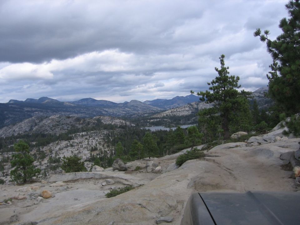 Dark gray vehicle hood in foreground, overlooking a vast mountain range with a lake in the distance under a cloudy sky
