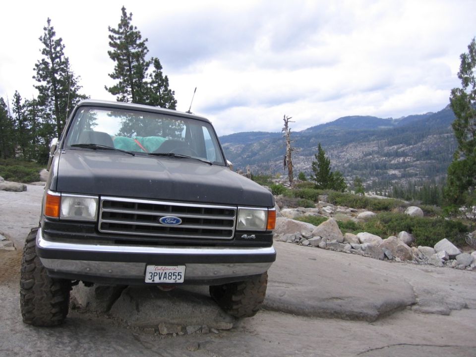 Dark gray Ford Bronco with off-road tires parked on rocks, mountains in background. License plate 3PVA855