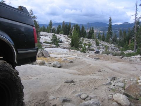 1989 Ford Bronco - Two vehicles navigate a rocky, off-road trail in a mountainous, wooded area under a cloudy sky.