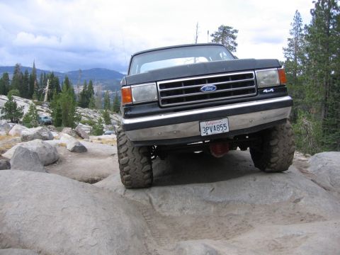1989 Ford Bronco - A dark-colored Ford truck navigates a rocky trail with trees and mountains in the background.