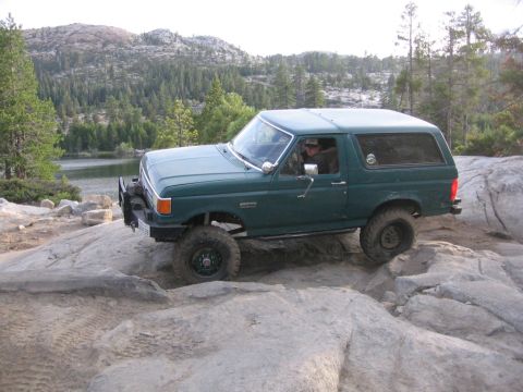 1989 Ford Bronco - This green Ford Bronco navigates a rocky terrain with a lake and trees in the background.