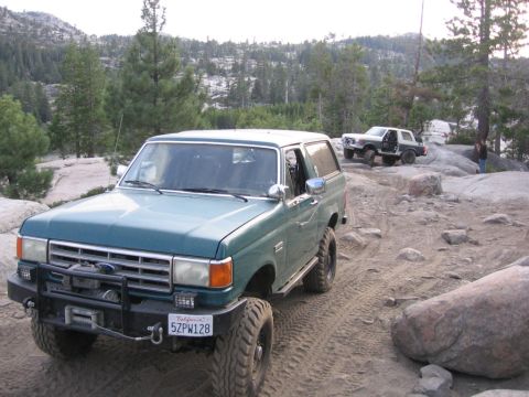 1989 Ford Bronco - Two Ford Broncos navigate a rocky, off-road trail with trees and mountains in the background.