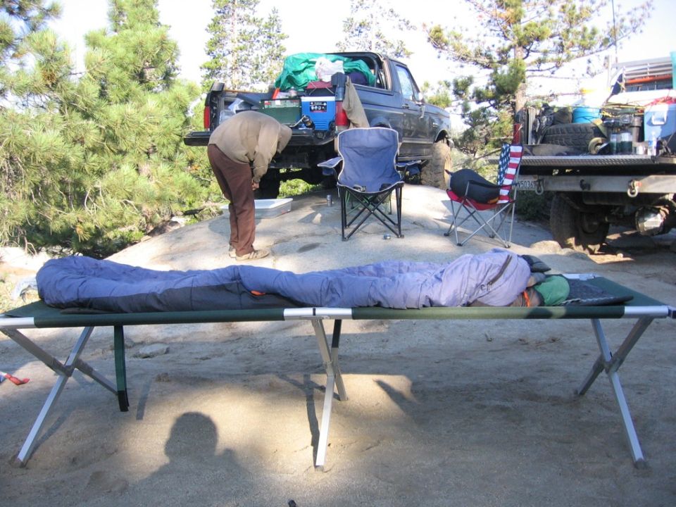 Two cots with sleeping bags are set up in front of two pickup trucks parked on a rocky, sandy area with pine trees