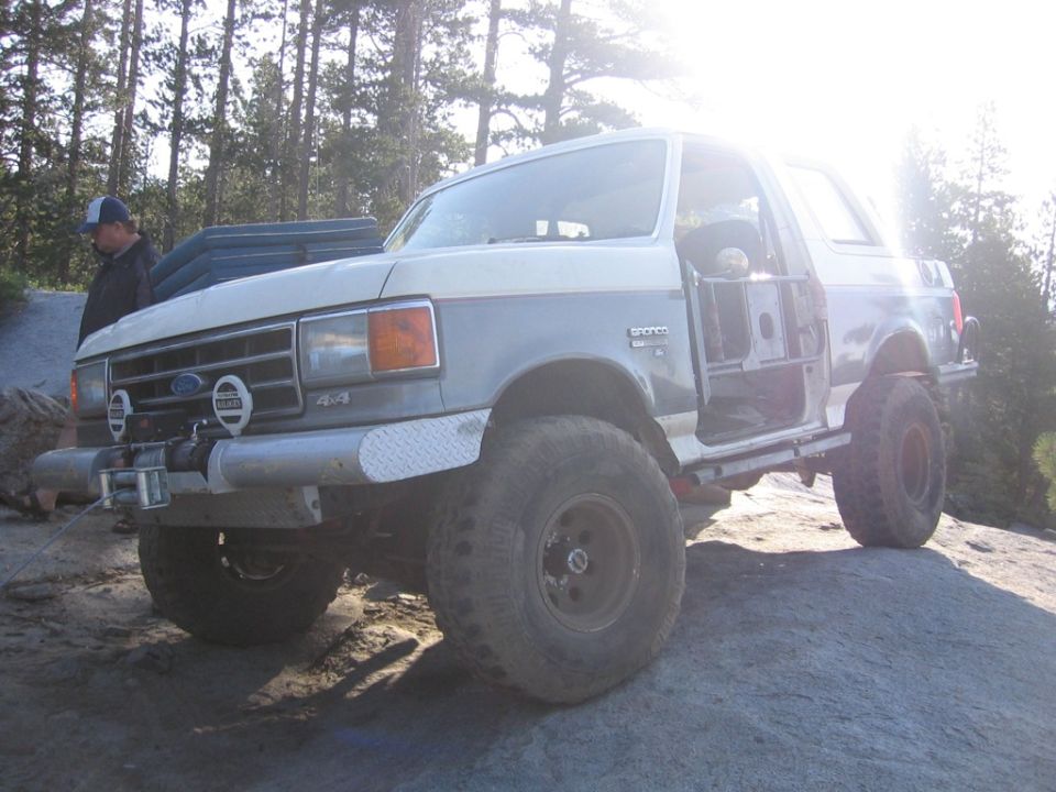 Two-tone Ford Bronco with doors removed, parked on rocks. A person stands nearby
