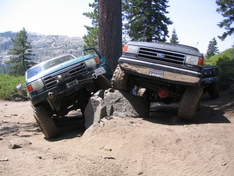 1989 Ford Bronco - Two Ford trucks are shown off-roading, with one climbing over rocks on a dirt trail.