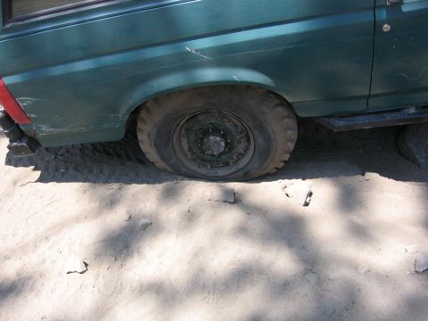 1989 Ford Bronco - The rear wheel of a green SUV is flat and resting on the ground in a sandy area.