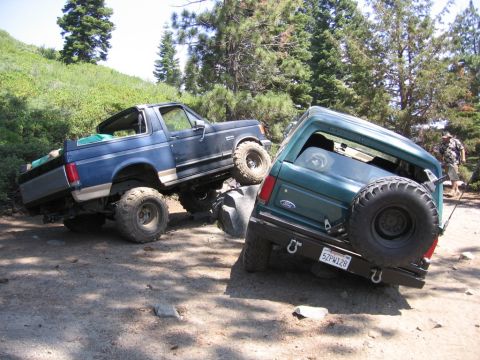 1989 Ford Bronco - Two Ford Broncos navigate a challenging off-road trail, one of them tilted precariously.