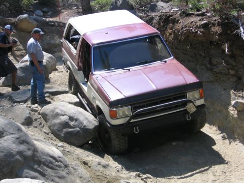 1989 Ford Bronco - A two-tone Ford Bronco is shown tackling a difficult off-road obstacle with two people observing.