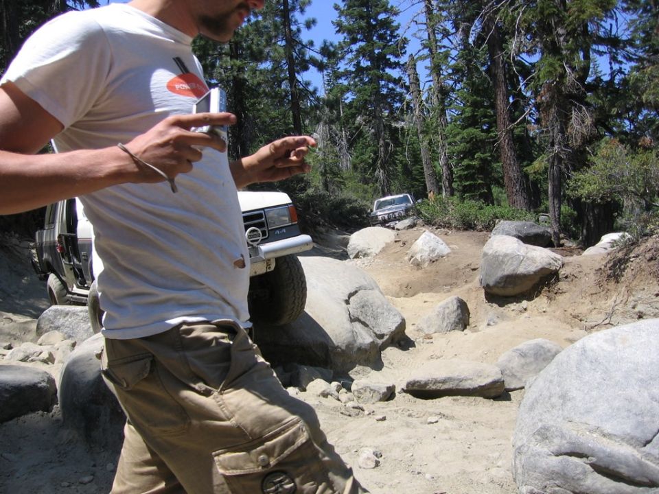 Two off-road vehicles navigate a rocky, wooded trail. A person in the foreground holds a phone