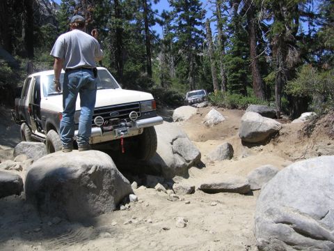 1989 Ford Bronco - A white and brown Ford Bronco navigates a challenging rocky trail with another vehicle visible in the background.