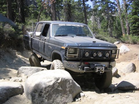 1989 Ford Bronco - This lifted, dark blue pickup truck navigates a rocky off-road trail, with "UNDER CONSTRUCTION 4X4" visible on the windshield.