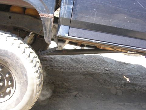 1989 Ford Bronco - The vehicle's undercarriage and large off-road tire are visible against a sandy, uneven terrain.
