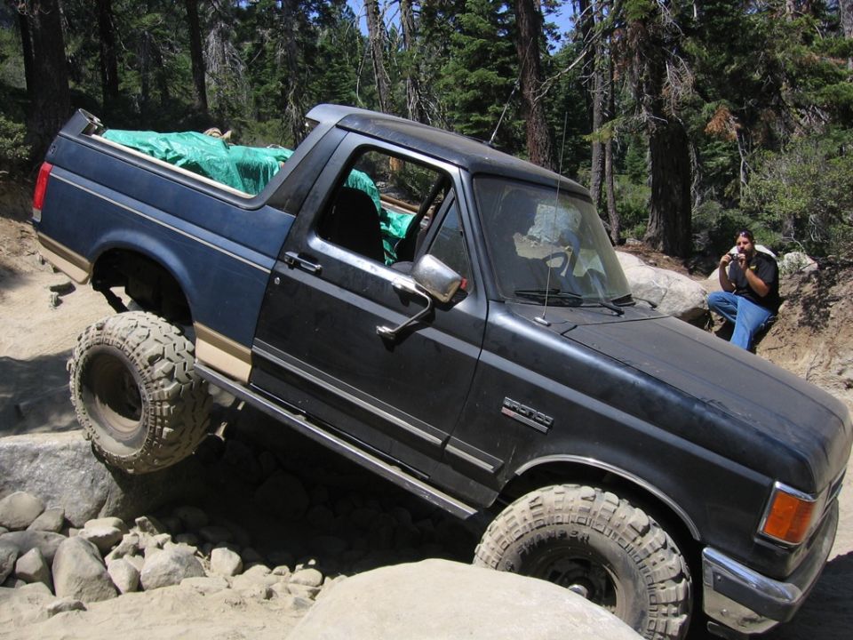 Dark blue and black Ford Bronco off-roading over rocks, with a person sitting in the background