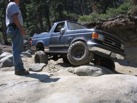 1989 Ford Bronco - A dark blue Ford Bronco navigates a challenging rocky trail with a person standing nearby.