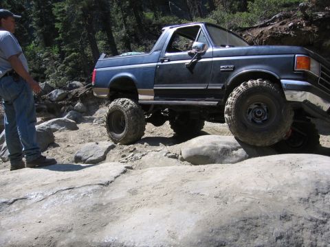 1989 Ford Bronco - A blue 1989 Ford Bronco is shown off-roading on a rocky trail with a person standing nearby.