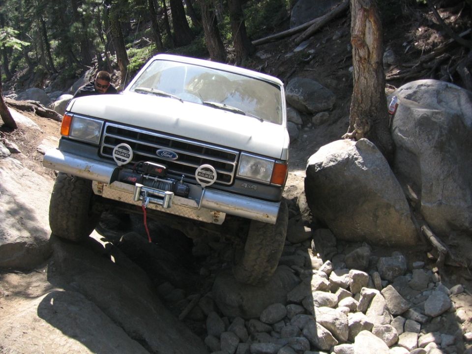 1989 Ford Bronco navigating a steep, rocky off-road trail with trees and large boulders