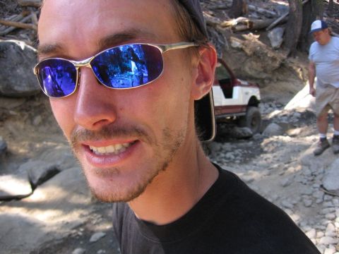 1989 Ford Bronco - A man wearing blue-tinted sunglasses smiles while an off-road vehicle and another person are visible on a rocky trail behind him.