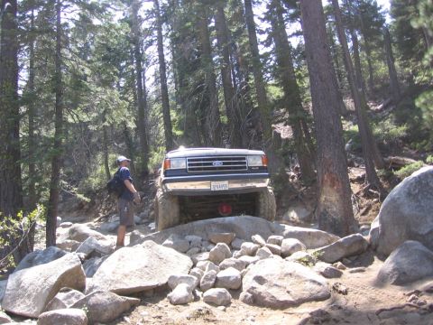 1989 Ford Bronco - A man stands by a lifted Ford Bronco navigating a challenging rocky terrain in a forest.