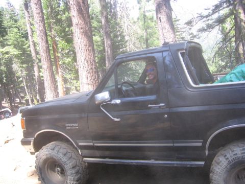 1989 Ford Bronco - A black Ford Bronco is parked off-road in a forest with a driver behind the wheel.