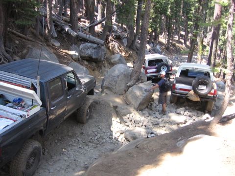 1989 Ford Bronco - Off-road vehicles tackle a challenging, boulder-strewn path in a forest.