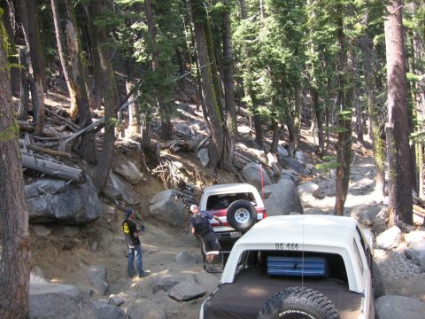 1989 Ford Bronco - Two SUVs navigate a challenging, rocky trail through a dense forest.