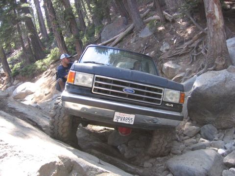 1989 Ford Bronco - A Ford Bronco is shown off-roading on a steep, rocky path surrounded by trees.