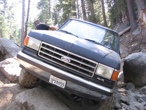 1989 Ford Bronco - A dark blue Ford Bronco navigates a challenging rocky terrain on a wooded trail.