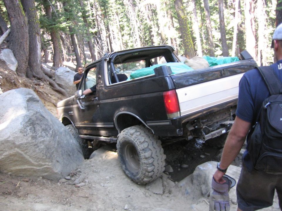 1989 Ford Bronco navigating a steep, rocky off-road trail with trees on either side