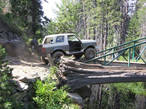 1989 Ford Bronco - A two-tone Ford Bronco navigates a challenging off-road obstacle, crossing a fallen log that spans a ravine.