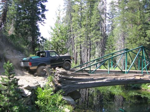 1989 Ford Bronco - A black Ford Bronco navigates a rough dirt path next to a green metal bridge crossing a body of water.