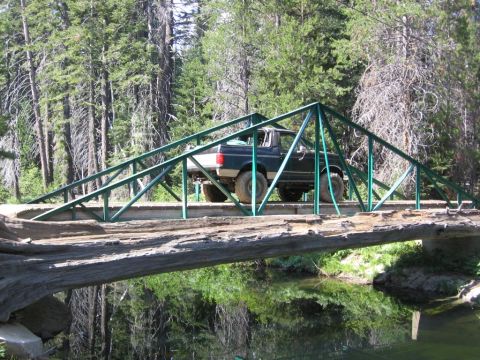 1989 Ford Bronco - A pickup truck drives across a rustic bridge in a forest setting.