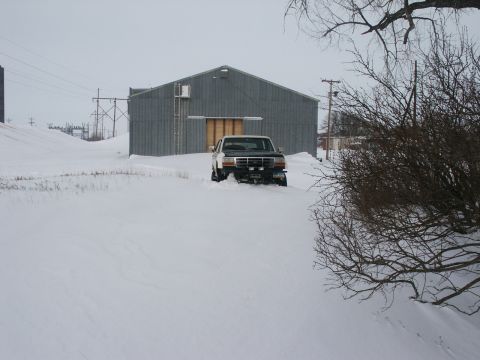 1990 Ford Bronco - A white and black Ford F-Series truck is partially buried in snow in front of a large metal building.