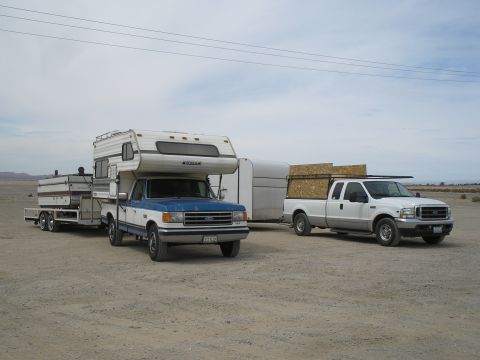 1989 Ford F-350 - A blue and white 1989 Ford F-350 pickup truck with a camper and trailer is parked next to a white pickup truck carrying lumber.
