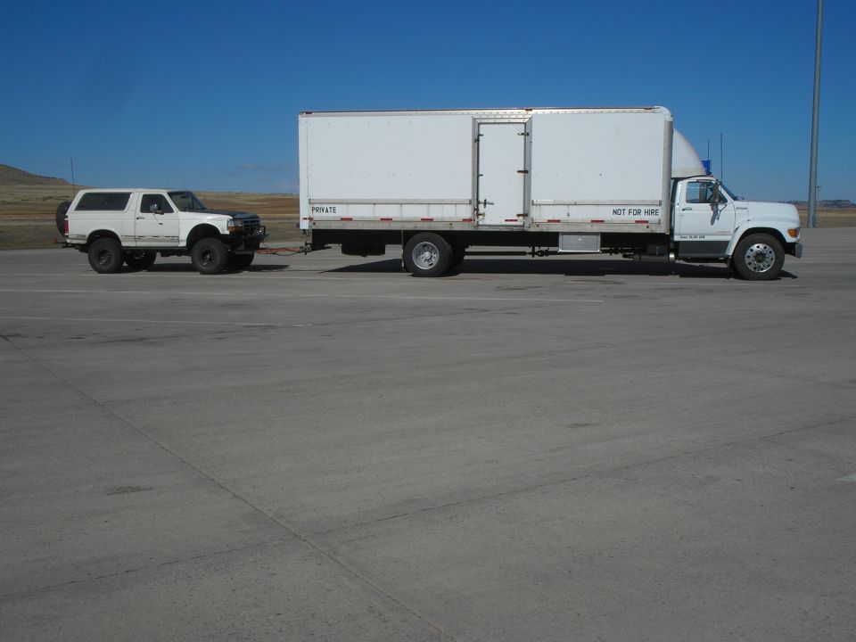 White SUV towing a white box truck on a paved lot under a clear blue sky