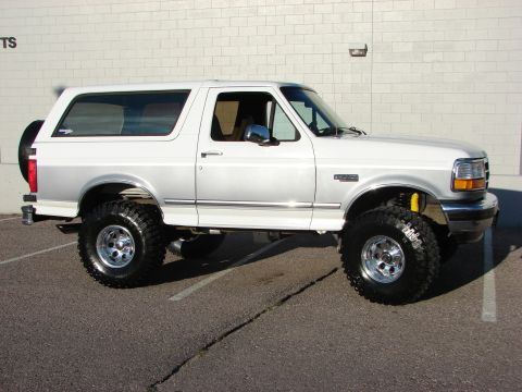 1995 Ford Bronco - Passenger-side profile of a white Ford Bronco two-door SUV lifted on mud-terrain tires with polished five-slot aluminum wheels, parked agai…